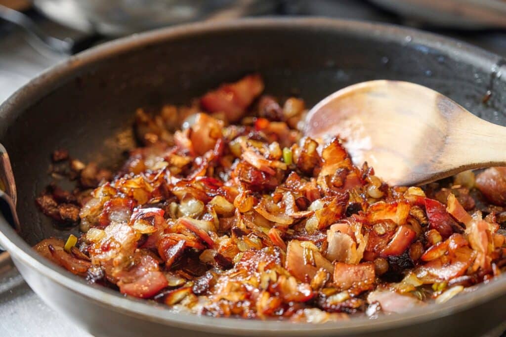 Bacon and caramelized onion cook slowly in a pan stirred with a wooden ladle in a traditional kitchen A rustic preparation that evokes home-cooked meals and authentic Mediterranean flavors | Aux Fourneaux Recette de pulled pork au four