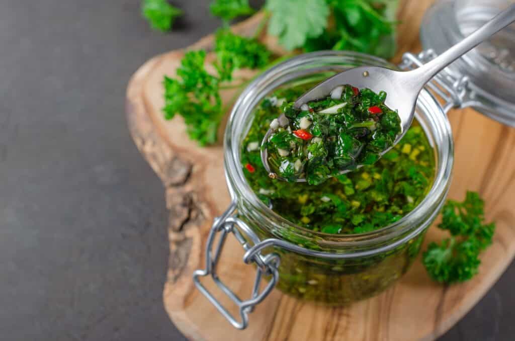 Traditional argentinian chimichurri sauce made of parsley cilantro garlic and chili pepper in a glass jar focus on a spoon with sauce Selective focus horizontal image | Aux Fourneaux La vraie sauce chimichurri traditionnelle