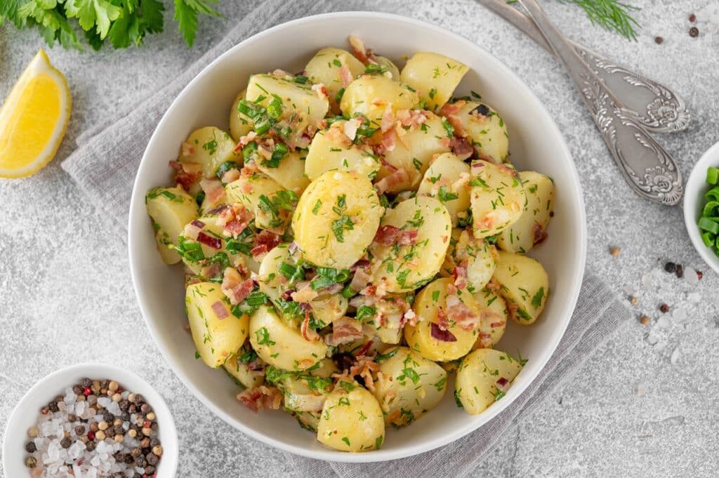 Warm potato salad with bacon onion fresh herbs and vinegar dressing in a bowl on a gray concrete background German Kartoffelsalat | Aux Fourneaux Salade de pommes de terre grand-mère traditionnelle