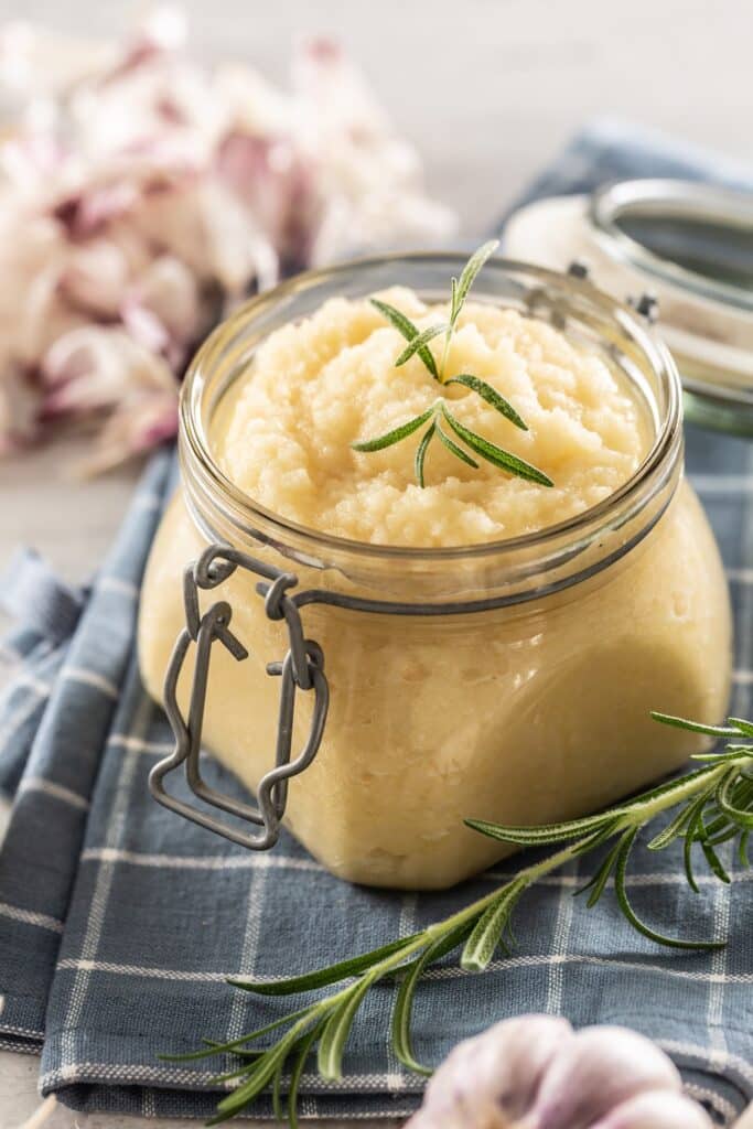 Aromatic garlic paste in a glass jar laid on rustic kitchen cloth with bulbs and peeled cloves and rosemary | Aux Fourneaux Purée d ail puissant anti-parasites recette naturelle et bienfaits