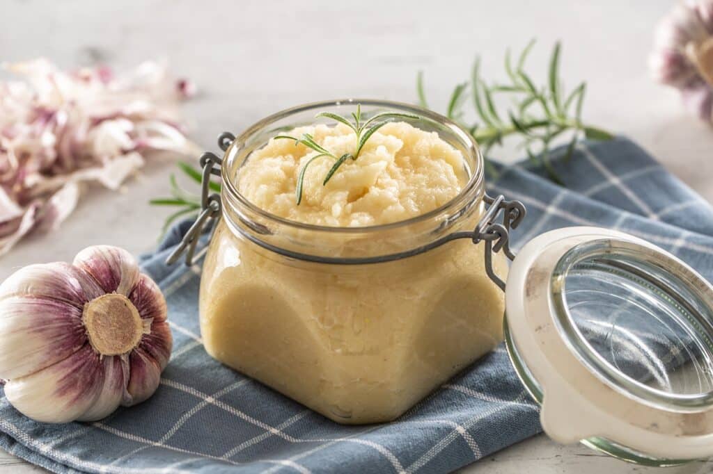 Aromatic garlic paste in a glass jar laid on rustic kitchen cloth with bulbs and peeled cloves and rosemary | Aux Fourneaux Purée d’ail puissant anti-parasites remede de grand mere