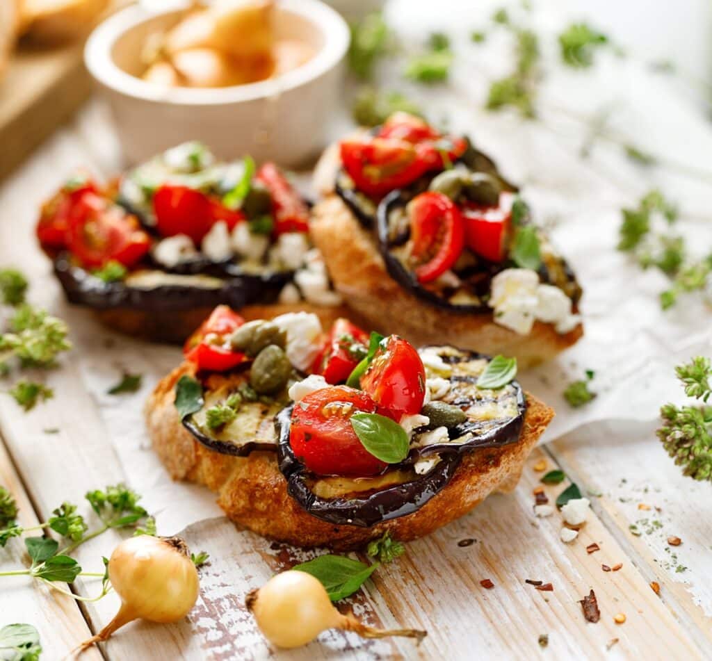 Bruschetta with grilled aubergine cherry tomatoes feta cheese capers and fresh aromatic herbs on a wooden table Delicious Mediterranean appetizer | Aux Fourneaux Bruschetta-style toasts topped with roasted eggplant, cherry tomatoes, crumbled cheese, and fresh basil on a rustic wooden board