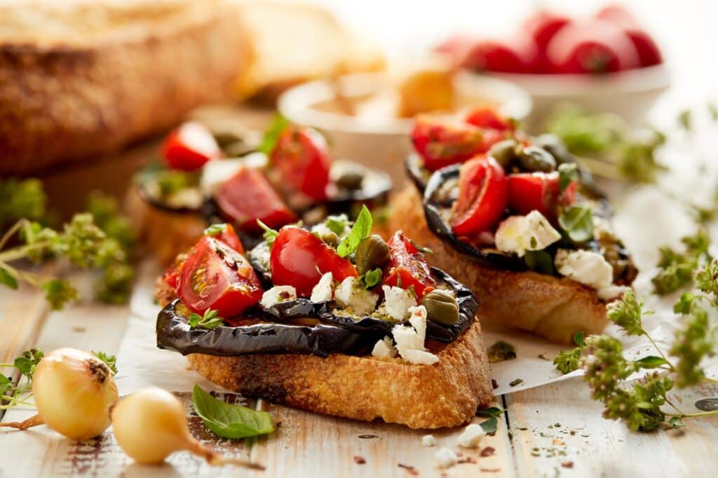 Toasts with grilled aubergine cherry tomatoes feta cheese capers and fresh aromatic herbs on a wooden table Delicious Mediterranean appetizer | Aux Fourneaux recette de bruschetta au four aux aubergines