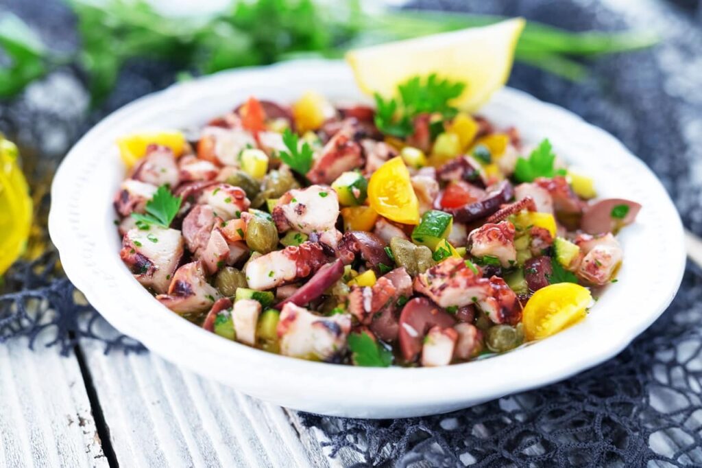 Fresh octopus salad | Aux Fourneaux Bowl of octopus salad with olives, peppers, corn, parsley, and a lemon wedge on the side.