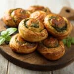 Stack of spinach and cheese puff pastry pinwheels on a wooden board, with basil garnish nearby.
