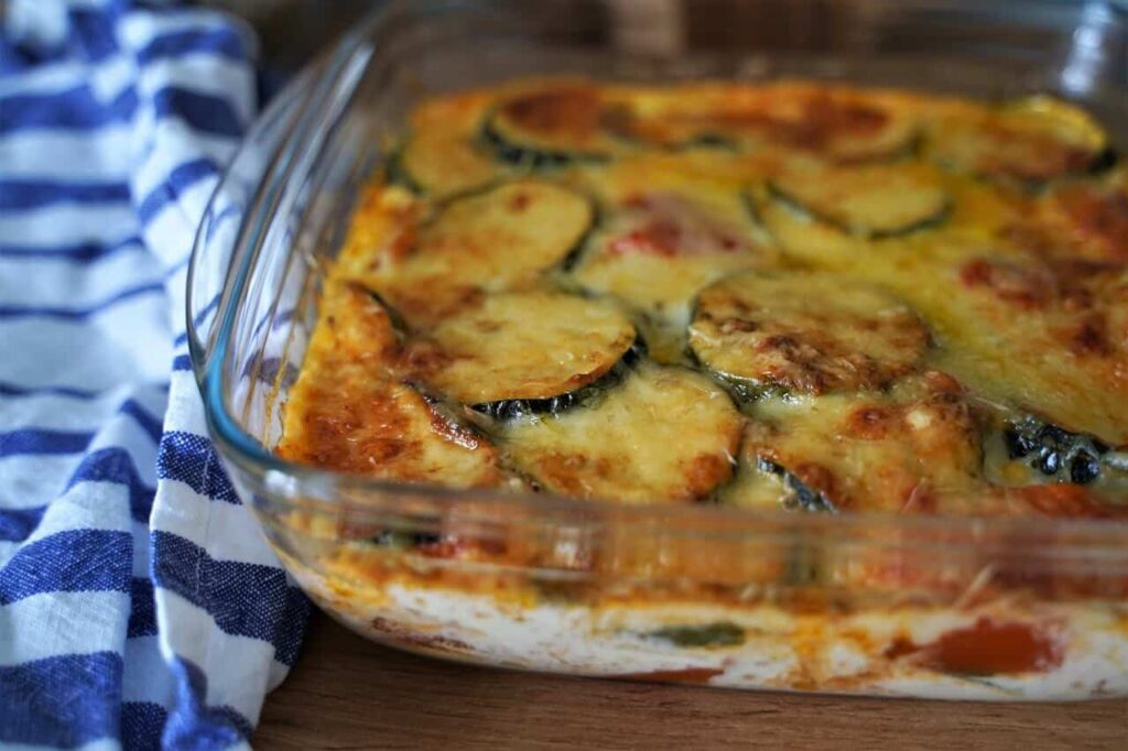 faut il éplucher les courgette pour faire un gratin | Aux Fourneaux Cheesy zucchini casserole bubbling in a clear glass baking dish on a wooden table with a blue-striped towel nearby.
