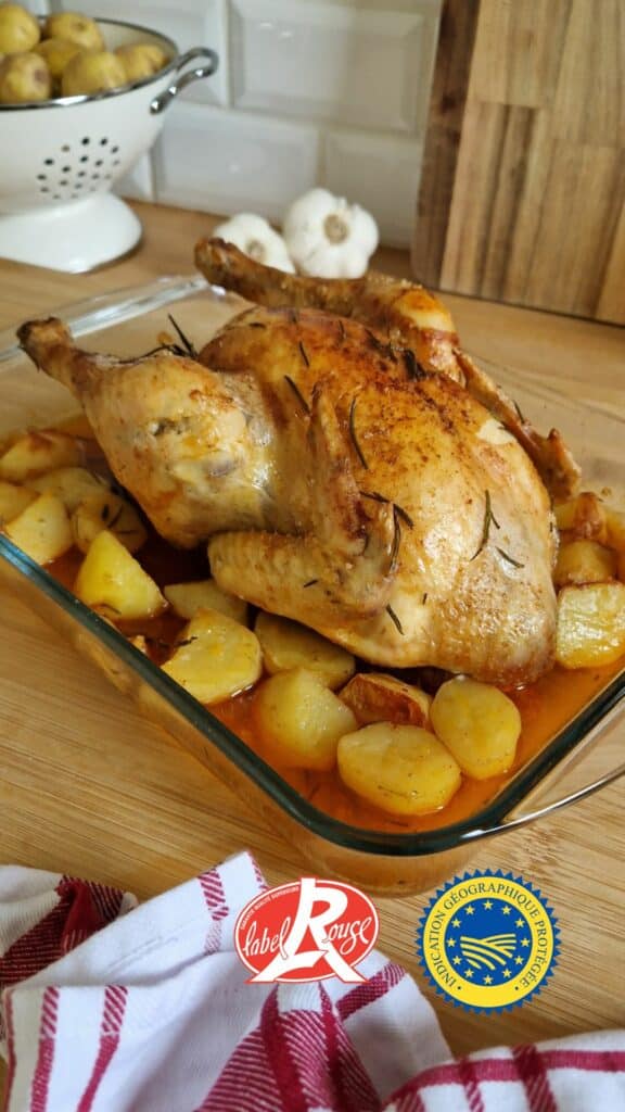 roti au four paprika pommes de terre | Aux Fourneaux Roasted whole chicken with crispy skin, rosemary sprigs, and potatoes in a glass baking dish on a wooden counter.